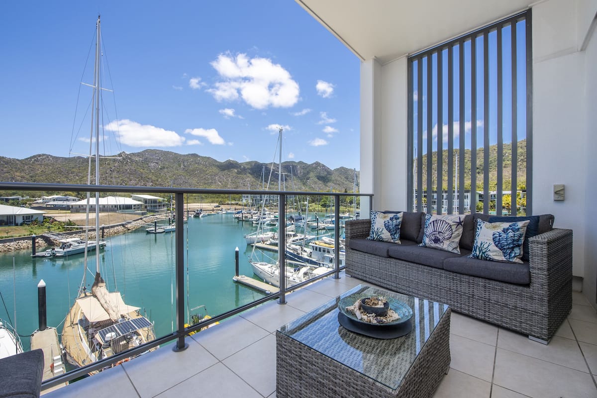 A balcony with a view of a marina, a wicker sofa, and a glass table.