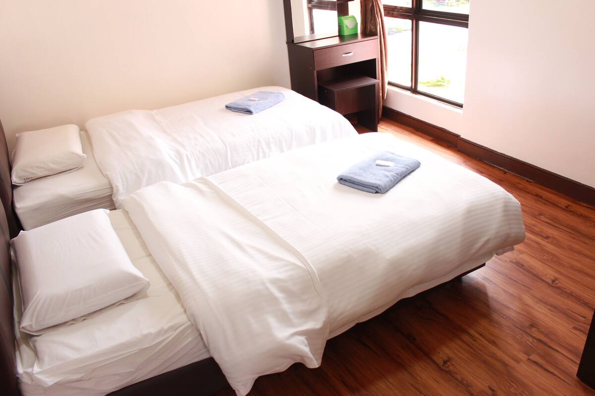 A double bed with white linens and a blue towel, a wooden nightstand, a mirror, and a window with curtains.