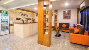 A reception area with a wooden reception desk, a refrigerator with beverages, a sofa with orange cushions, and a wall with a framed picture and a clock. - Phuthan Hotel (Tha Chang)