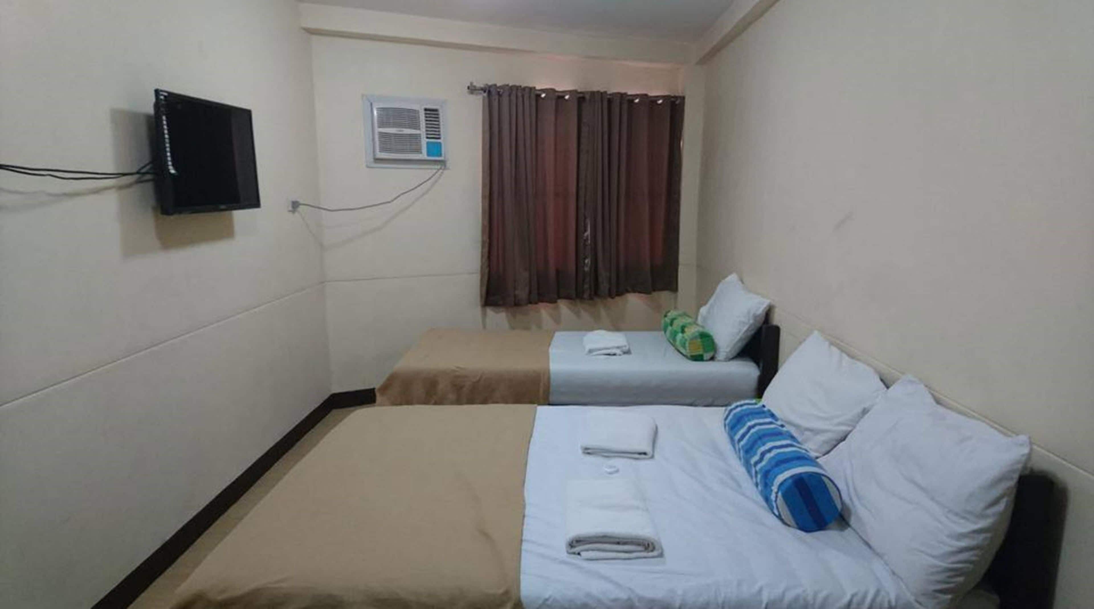 A small hotel room with two beds, a TV, an air conditioning unit, and a window with curtains.
