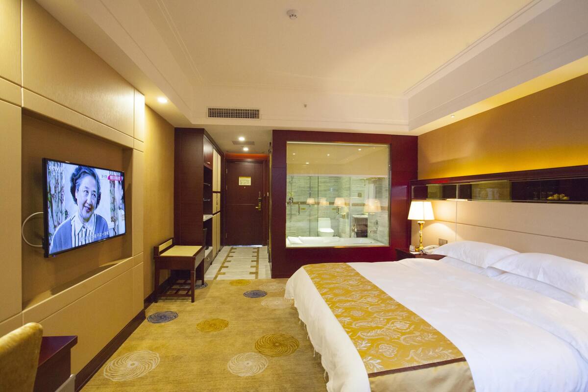 Une chambre d’hôtel avec un lit, un téléviseur fixé au mur, un bureau et une salle de bain visible à travers une porte en verre.