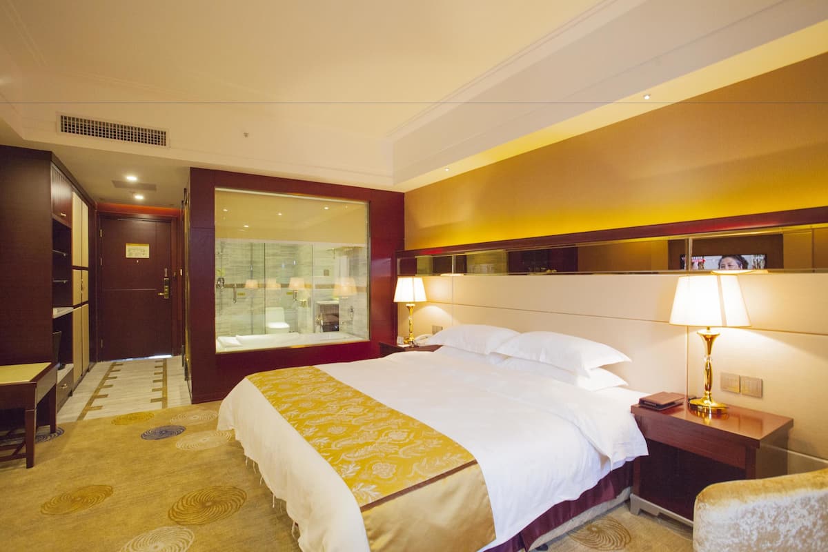 Une chambre d’hôtel avec un grand lit, des tables de chevet, un miroir et une salle de bain visible à travers une porte en verre.
