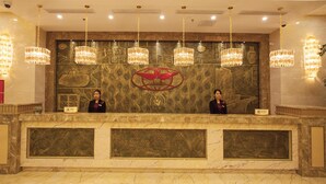 A reception desk with two staff members, a marble counter, and a wall with a logo and decorative panels. - Waijing Gloria Grand Hotel Anhui (Wuhu)