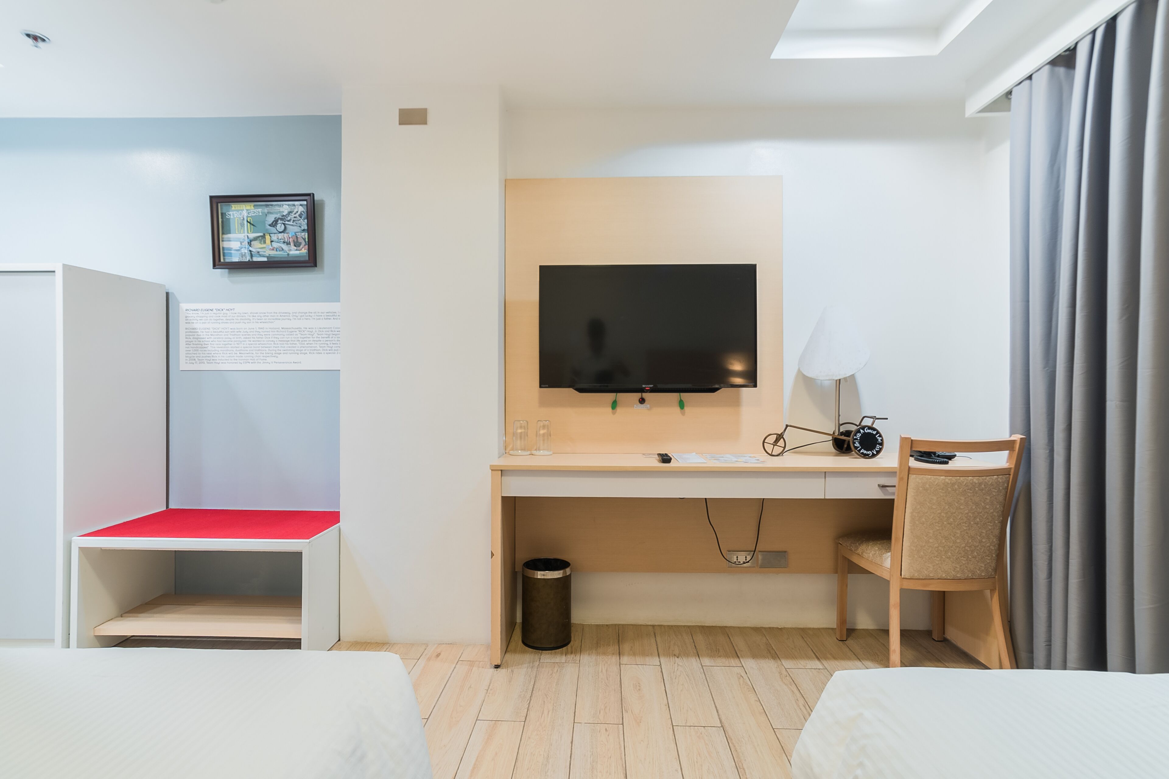 A hotel room with a bed, a desk with a TV, a chair, a small trash bin, and a red bench.