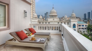 A balcony with two lounge chairs, a small table with a hat and magazines, and a view of a cityscape with domed buildings. - Maison De Camille Boutique Hotel (Ho Chi Minh City)