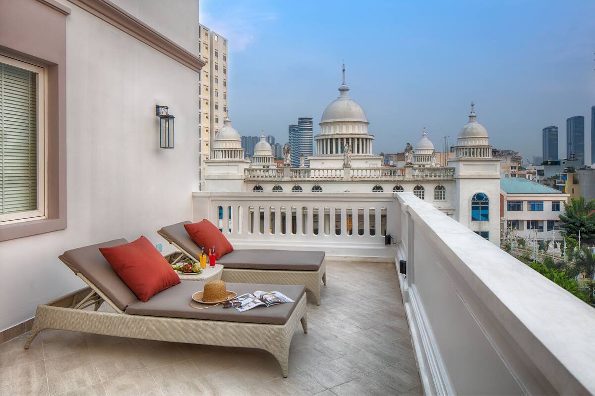 Ein Balkon mit zwei Loungestühlen, einem kleinen Tisch mit Hut und Zeitschriften sowie Blick auf eine Skyline mit Kuppelbauten.
