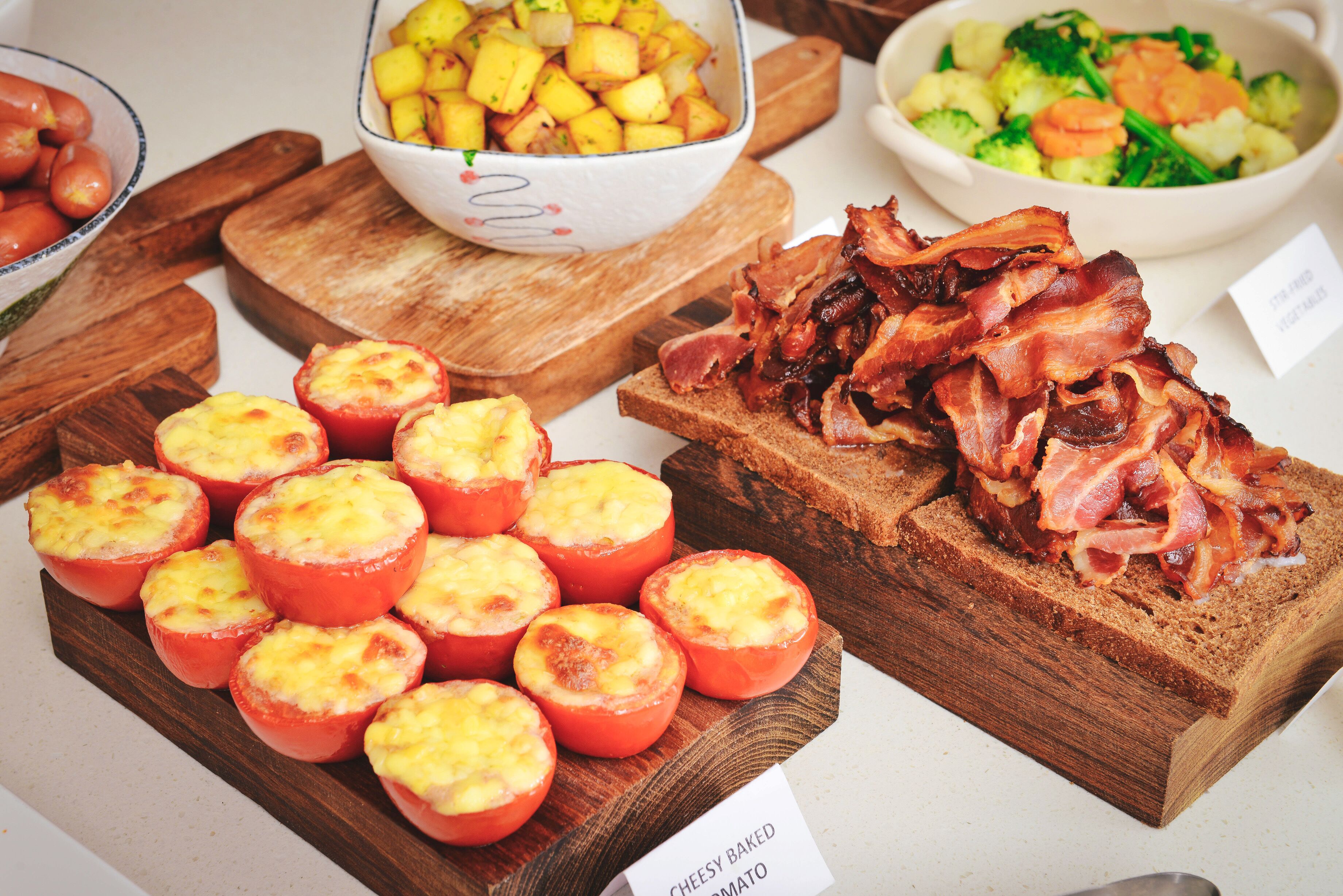 A wooden board with baked tomatoes and bacon, a bowl of potatoes, and a bowl of mixed vegetables.