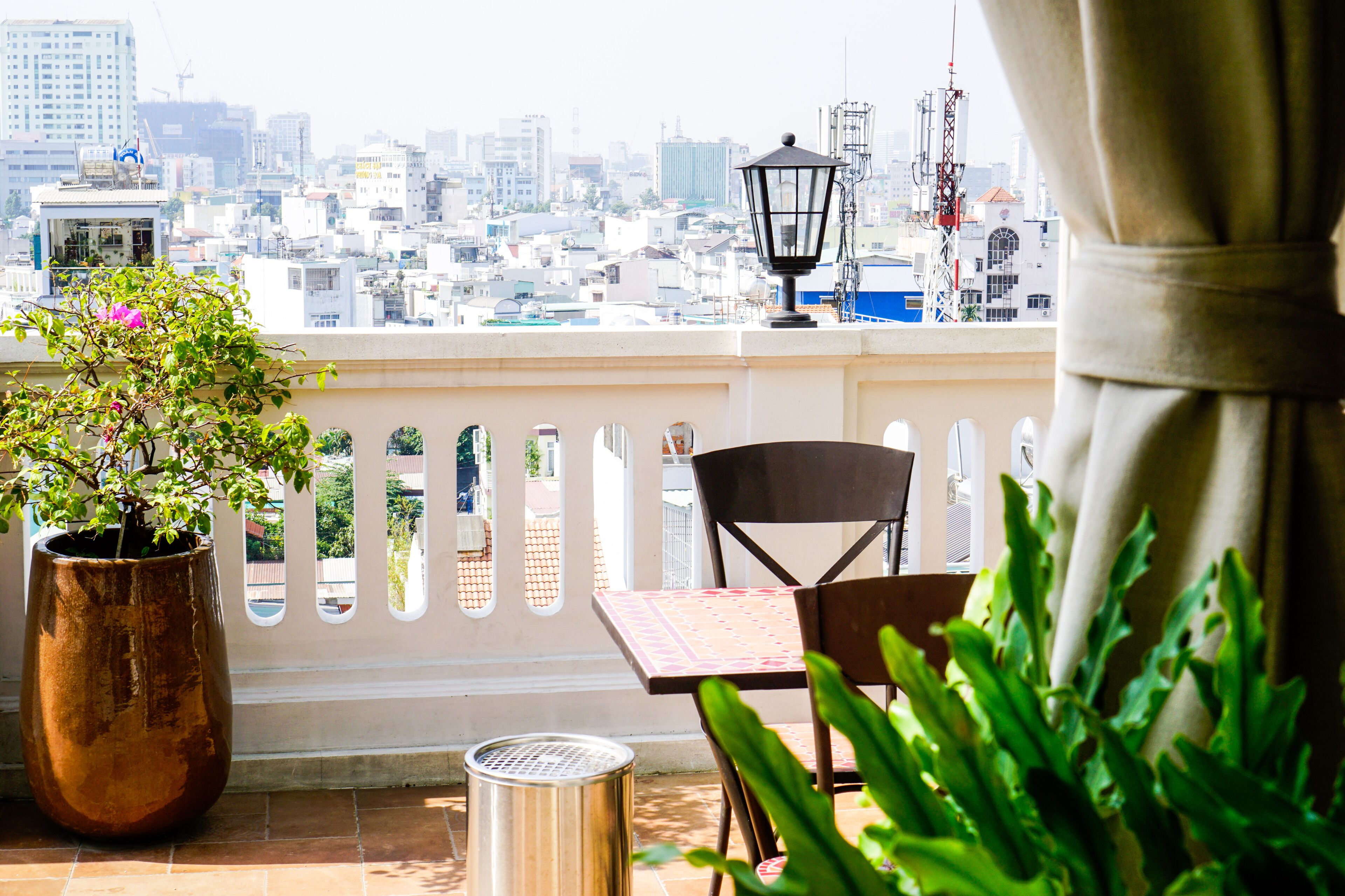 A balcony with a view of a cityscape, a potted plant, a trash can, and a table with a chair.