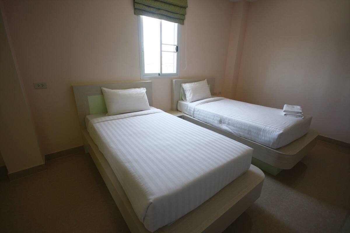 Two single beds with white bedding, a window with a green valance, and a plain wall.