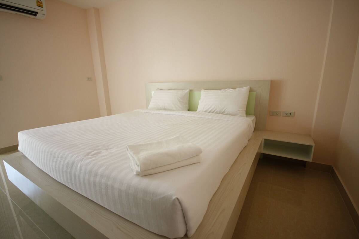 A neatly made bed with white linens and pillows, a bedside table, and a wall-mounted air conditioning unit.
