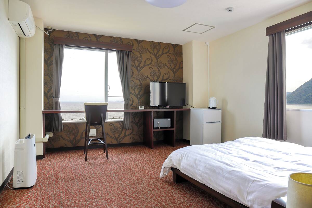 A hotel room with a bed, a desk with a computer, a chair, a small refrigerator, and a window with curtains.