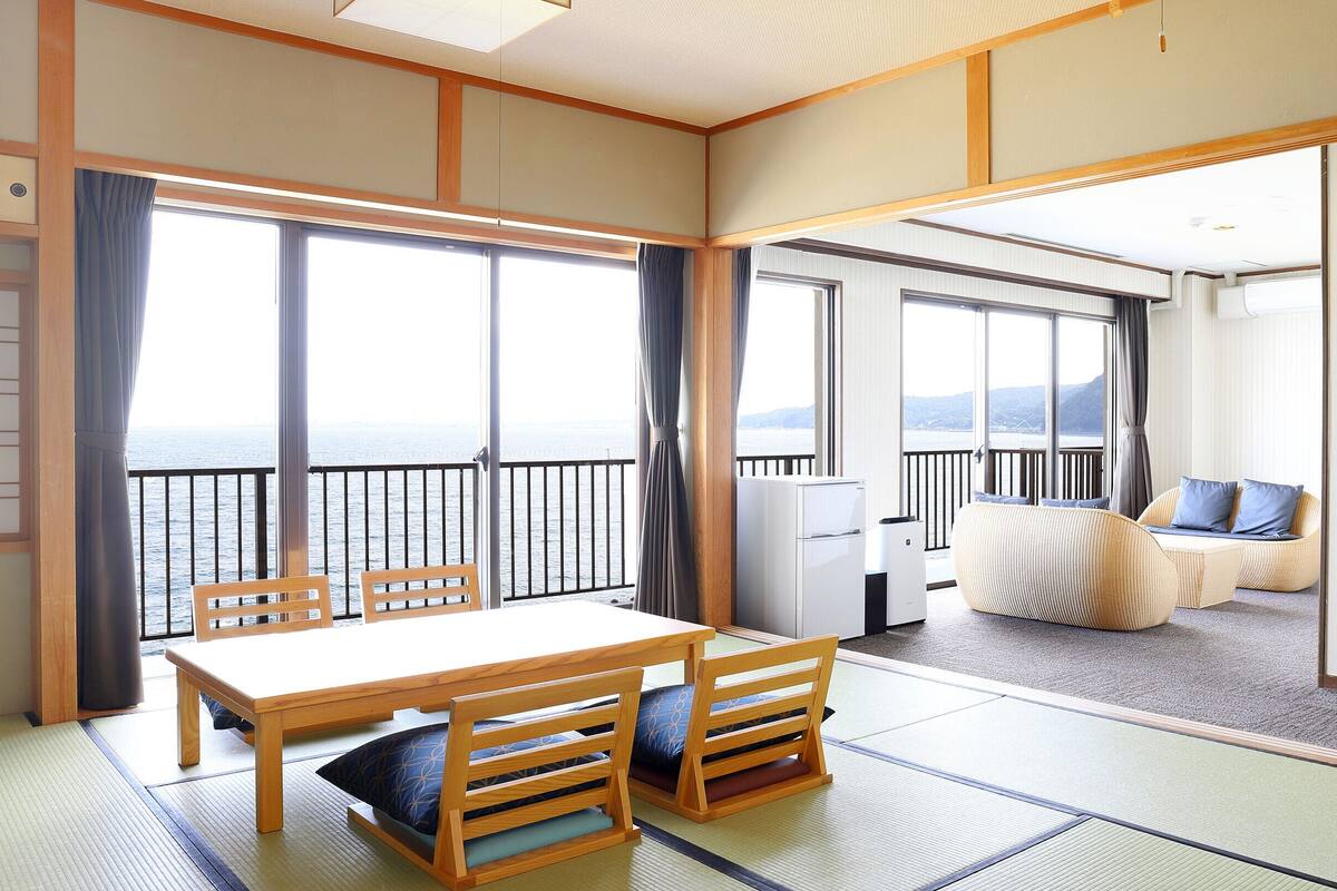 A traditional Japanese-style room with tatami flooring, a low table with chairs, a sofa with cushions, and a view of the sea through large windows.