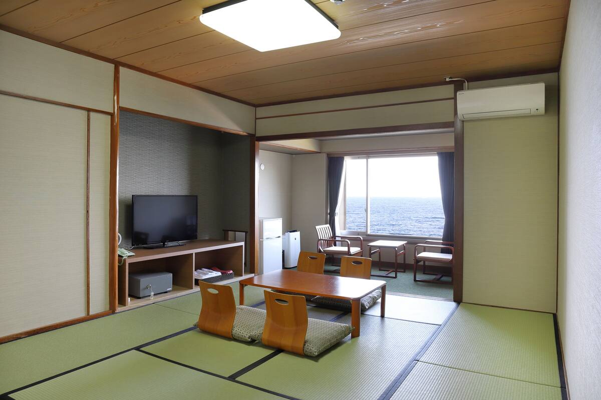 A room with tatami flooring, a television, a dining table with chairs, and a view of the sea through a window.