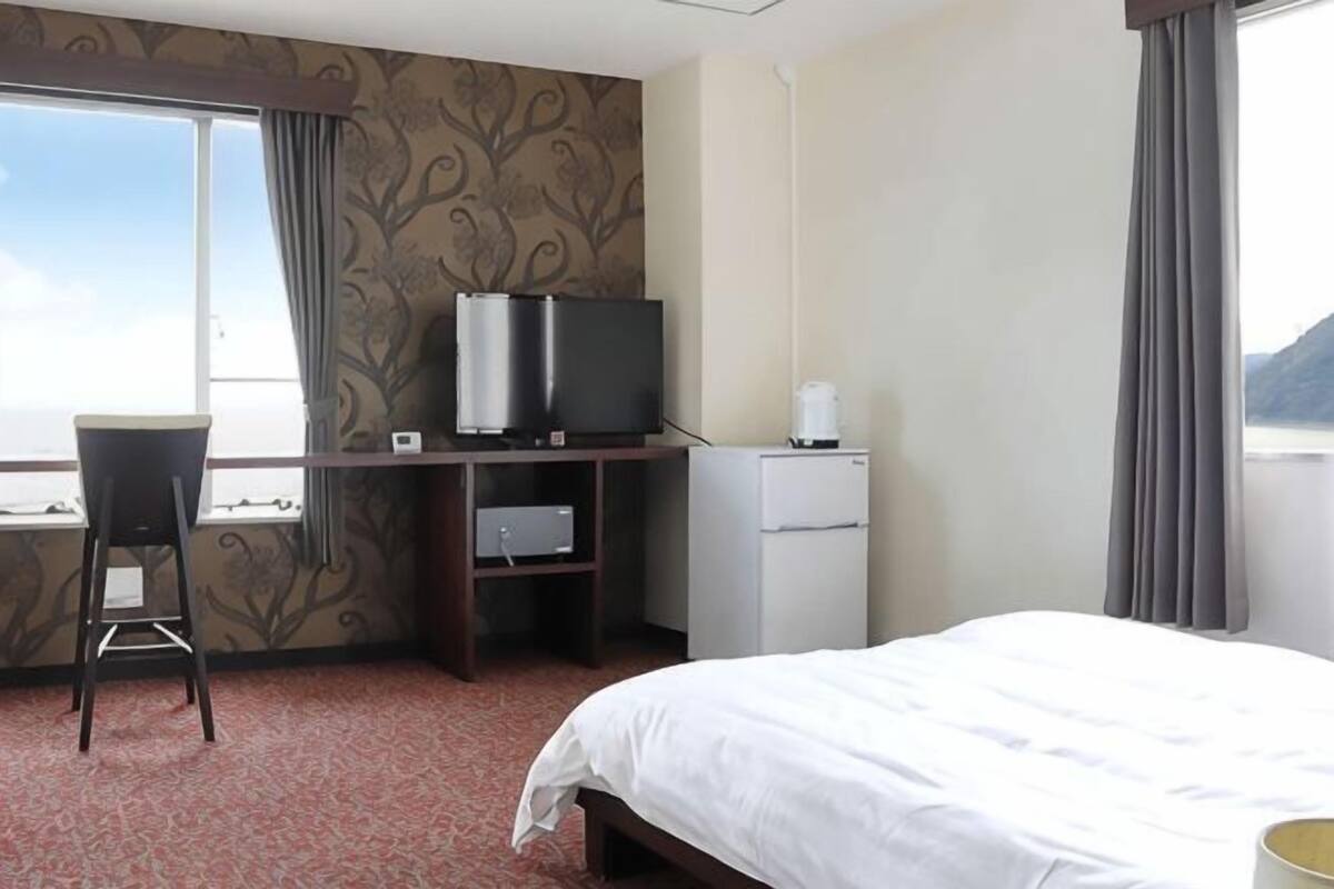 A hotel room with a bed, a desk with a chair, a television, a refrigerator, and a window with curtains.