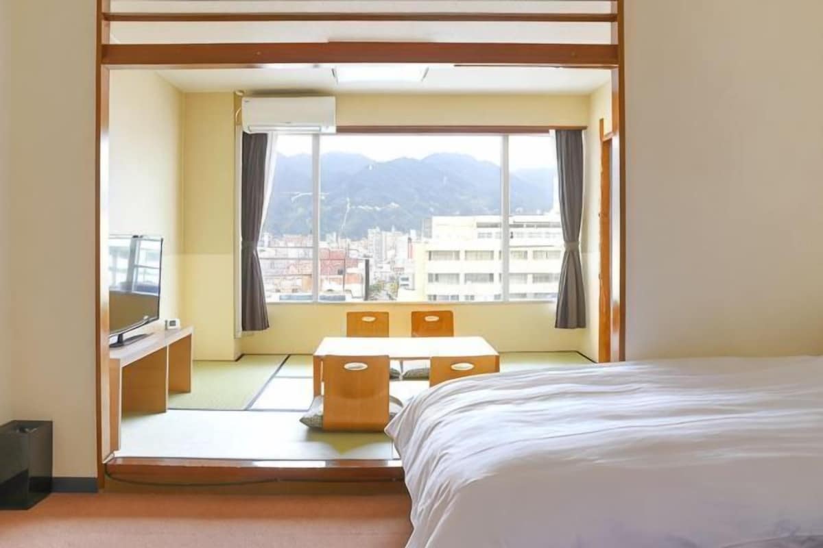 Una habitación con cama, escritorio, silla y ventana con vistas a edificios y montañas.