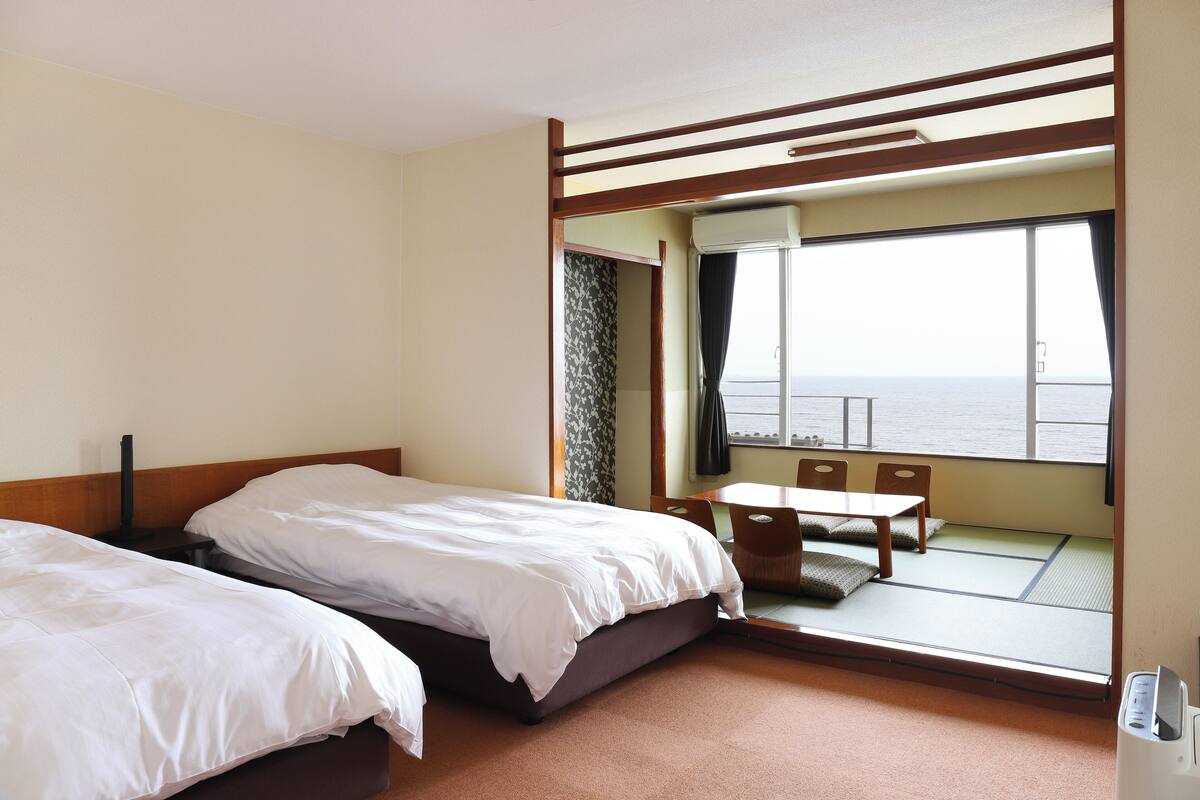 A hotel room with two beds, a small table, and a view of the sea through a large window.