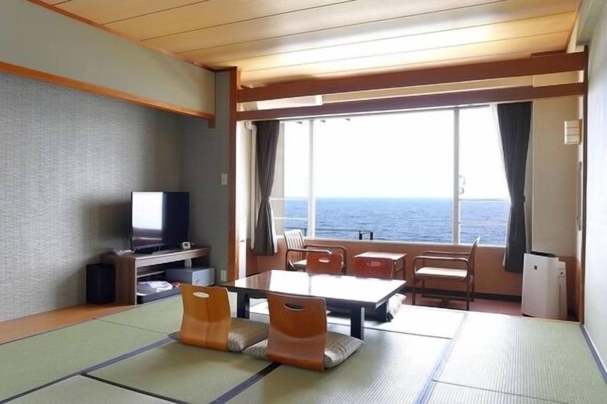 A room with tatami mats, a television, a table, chairs, and a view of the sea.