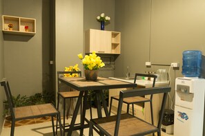 A compact kitchen with a dining table, chairs, a water dispenser, and a small shelf with decorative items. - Iris hostel (Chiang Mai)