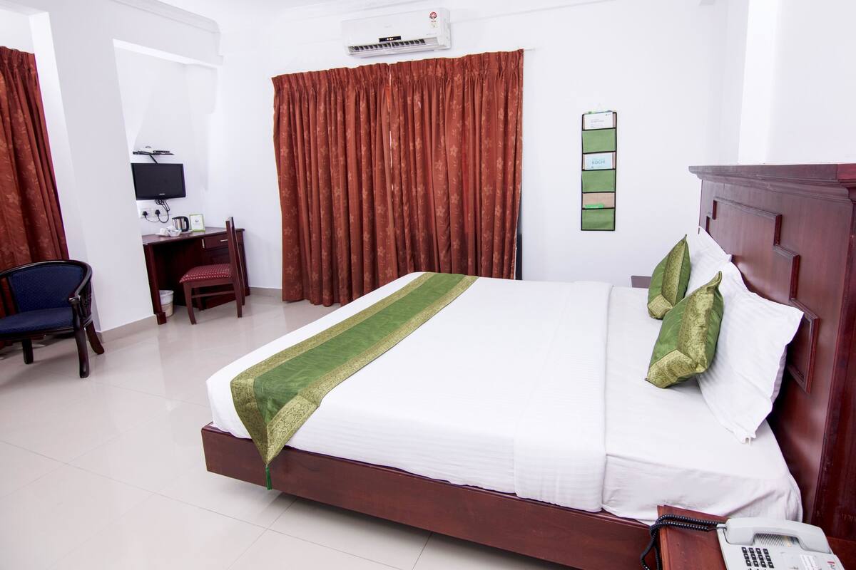 A hotel room with a bed, a desk with a chair, a television, and a wall-mounted air conditioner.