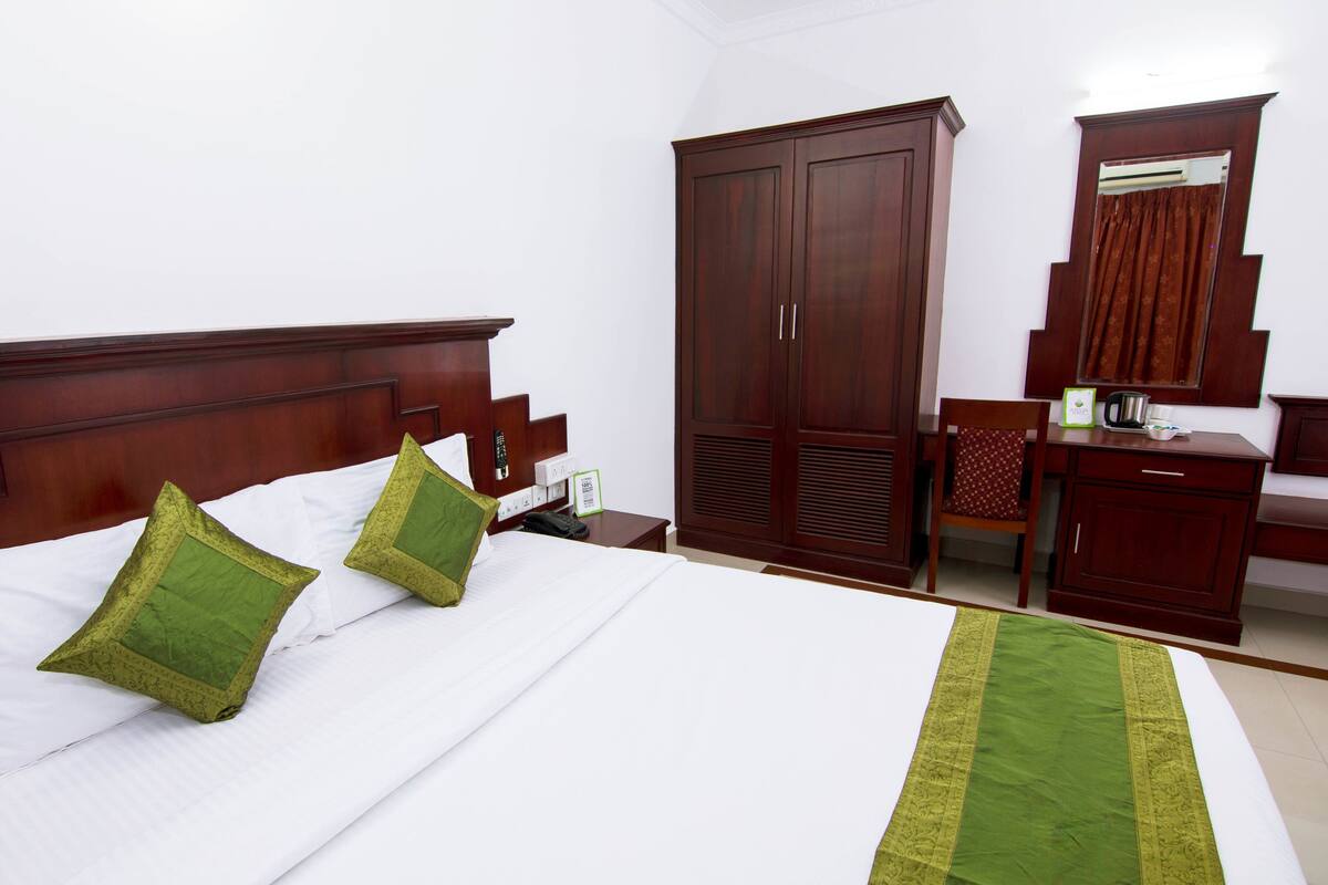 A neatly made bed with white linens and two green pillows, a wooden wardrobe, a small desk with a chair, and a mirror above the mirror.