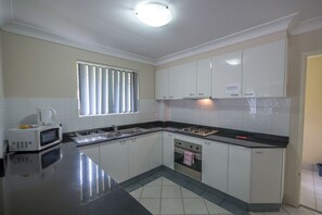 A modern kitchen with white cabinets, a black countertop, a built-in oven, and a window with blinds. - Parkside Apartments (Parramatta)