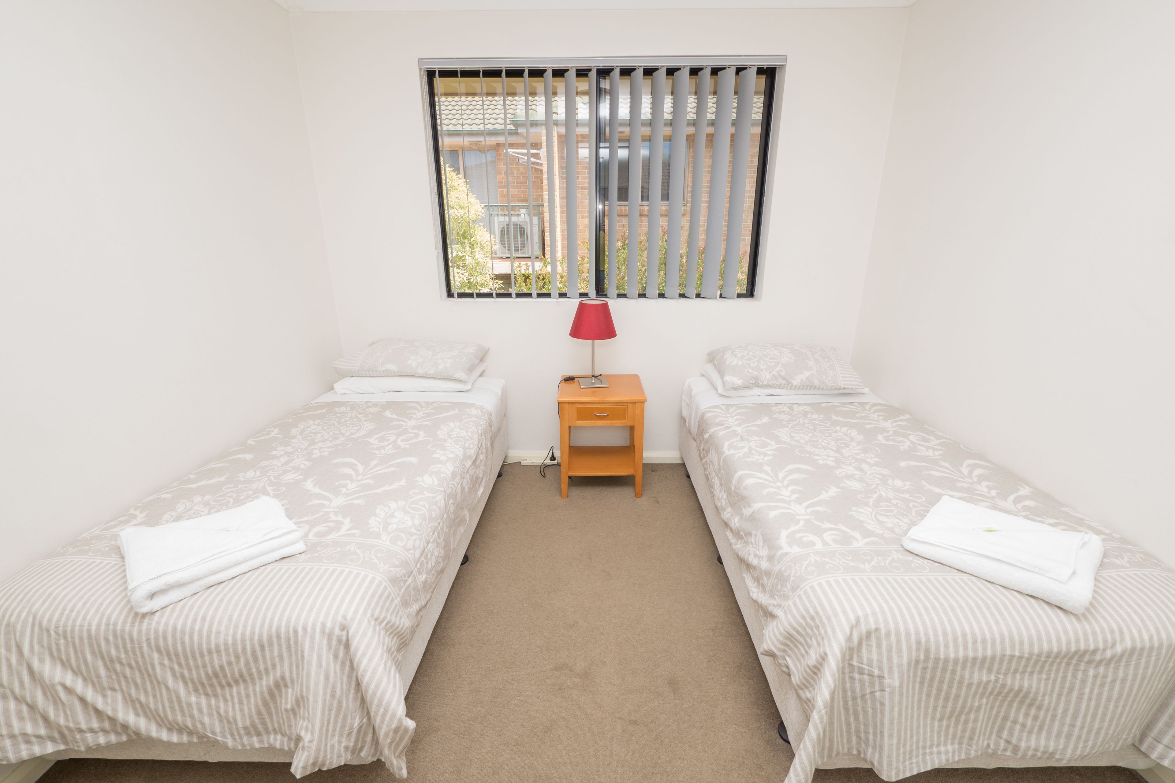 Un dormitorio con dos camas individuales, una mesita de noche de madera con una lámpara roja y una ventana con persianas verticales.
