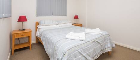 Una cama bien hecha con colcha a rayas, dos mesitas de noche con lámparas y una ventana con persianas.