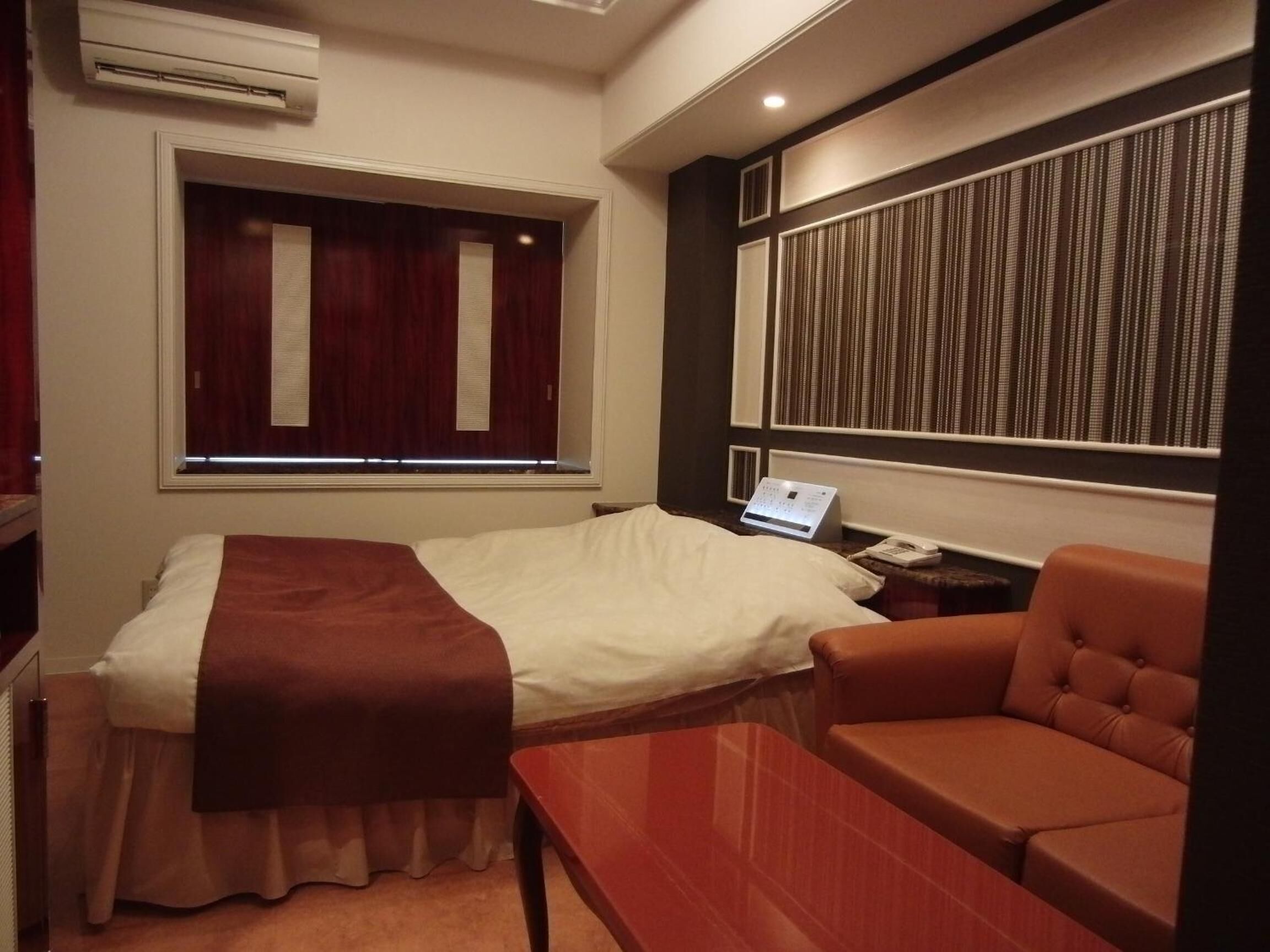 A hotel room with a bed, a bedside table with a clock, a chair, a window with curtains, and a wall panel.