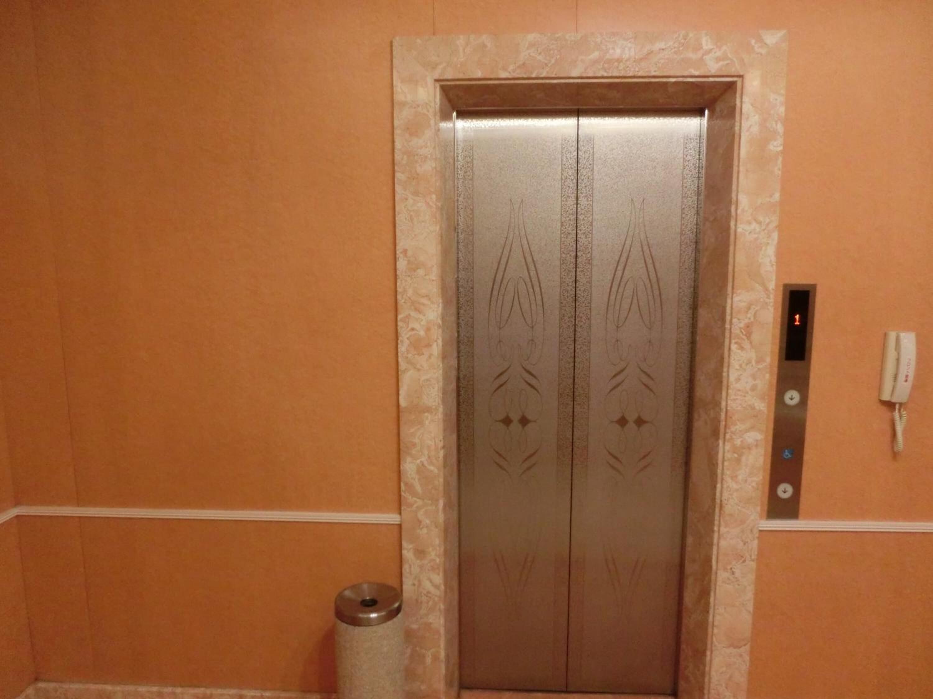 An elevator with a metallic finish and decorative patterns, a wall-mounted phone, and a trash can.
