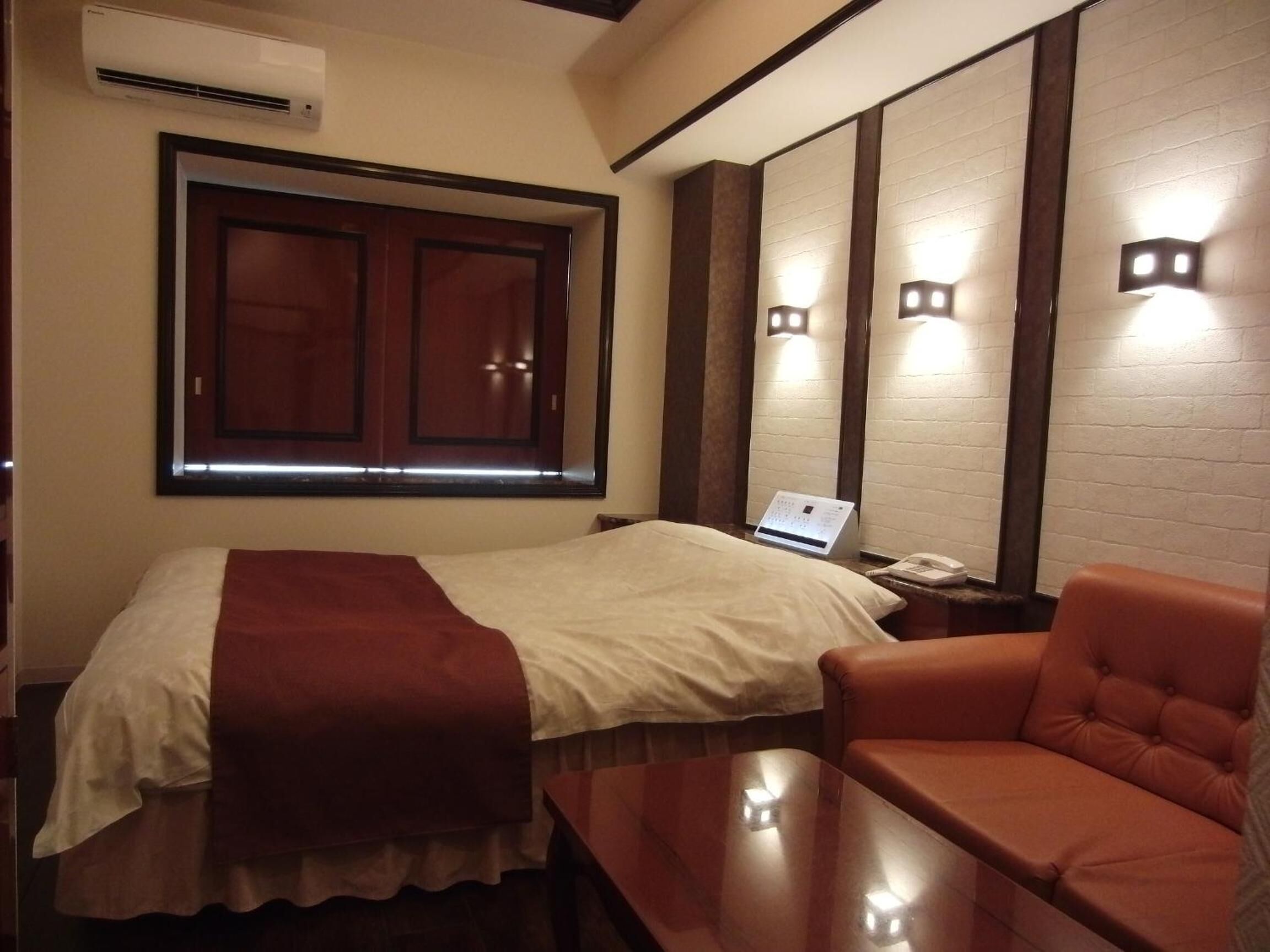 A hotel room with a bed, a laptop on a bedside table, a chair, and a wall-mounted air conditioner.