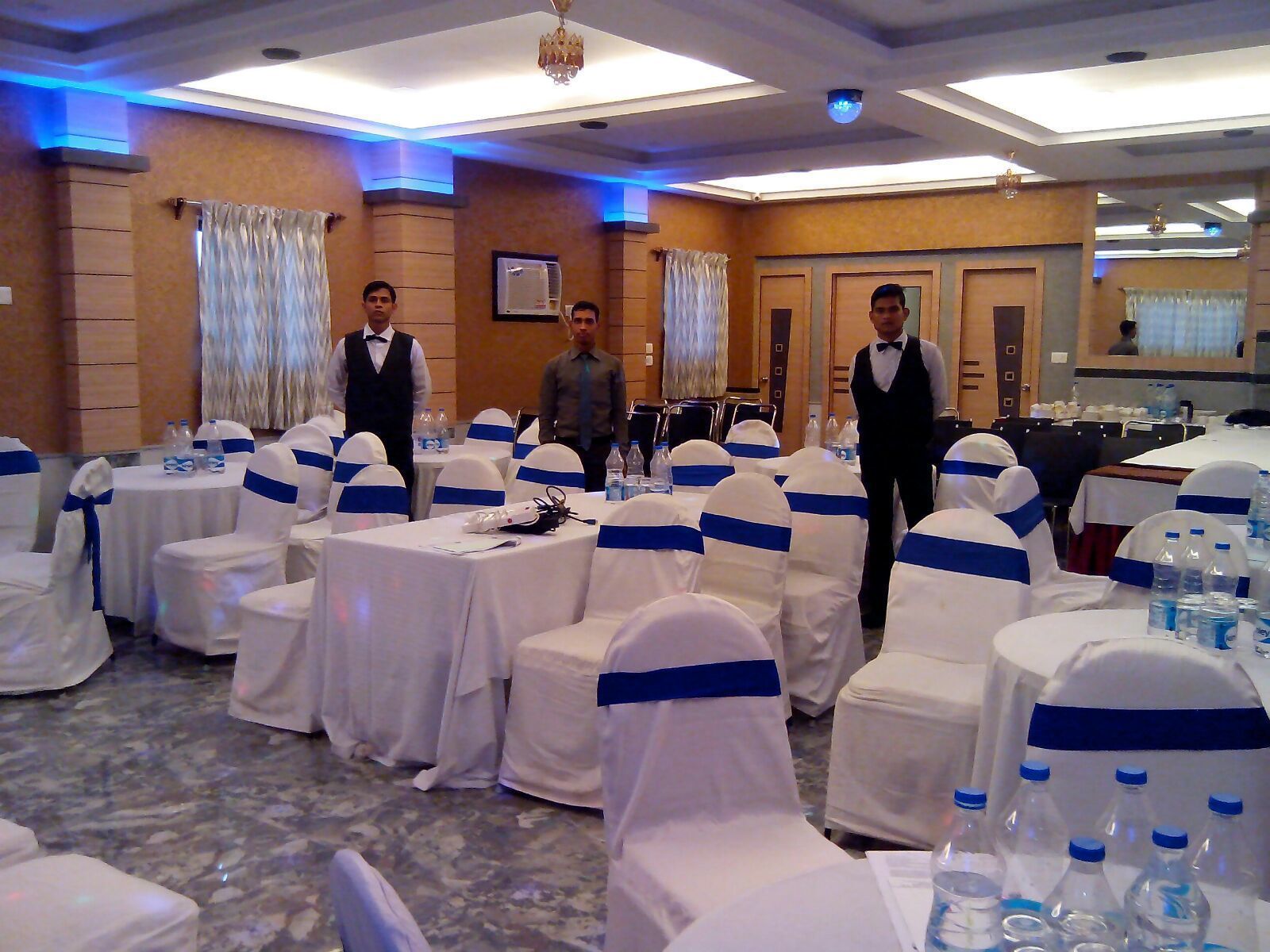 A banquet hall with round tables covered in white tablecloths and blue sashes, with waitstaff in the background.