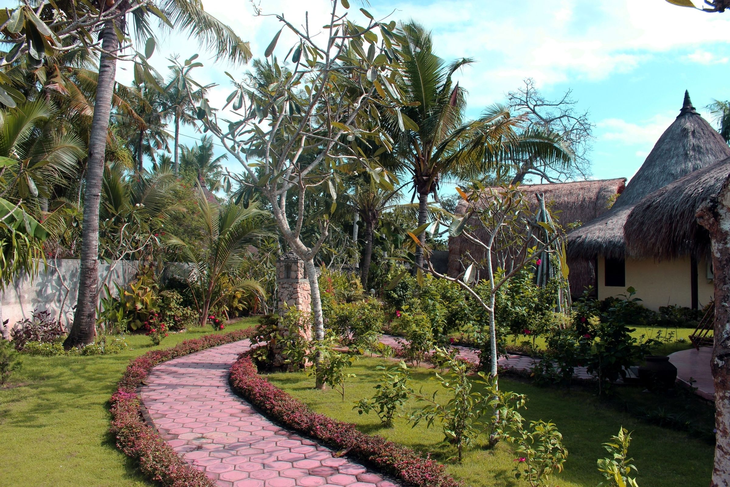A garden pathway leading to a thatched-roof building surrounded by palm trees and lush greenery.