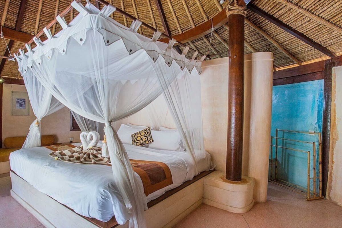 Een bed met hemelbed, voorzien van een witte hemel, een houten hoofdeinde en een decoratief kussen.