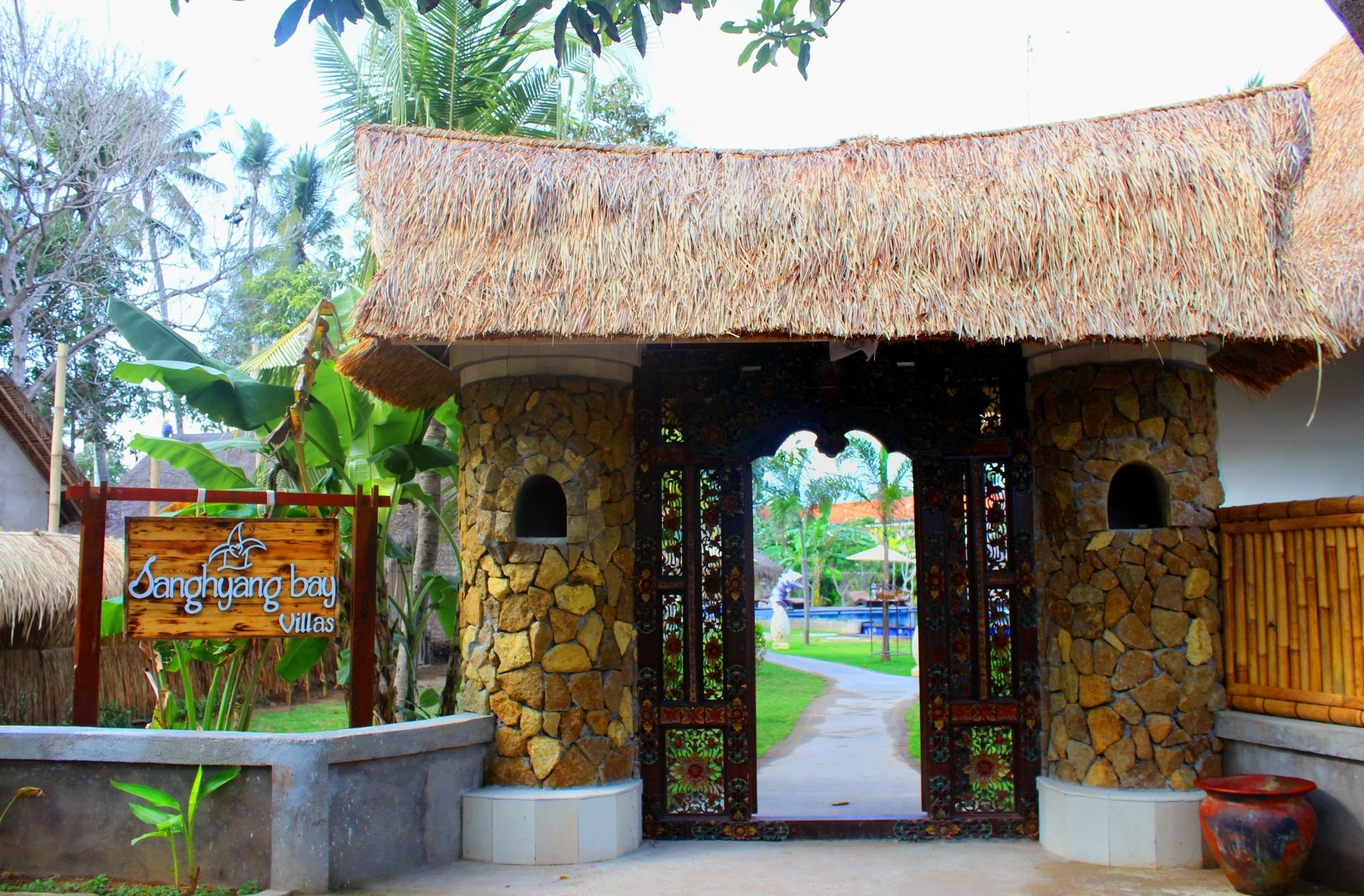 Una entrada con arco de piedra y techo de paja que conduce a un edificio con el letrero 'Sanghyang Bay Villas'.