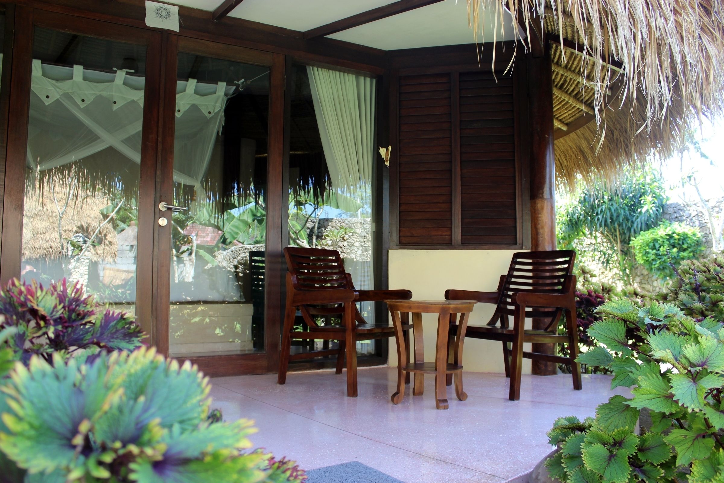 A covered patio area with two wooden chairs and a small table, surrounded by lush greenery and a thatched roof.
