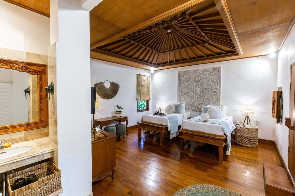 Una habitación con techo de madera, cama con ropa blanca, mesita de noche, lámpara, espejo y una obra de arte colgada en la pared.