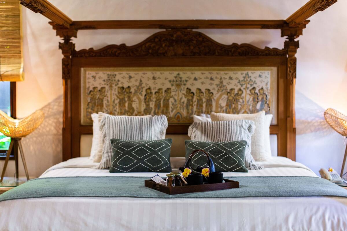 Una cama con cabecera de madera, dos almohadas decorativas, una bandeja con objetos y una pared con un tapiz estampado.