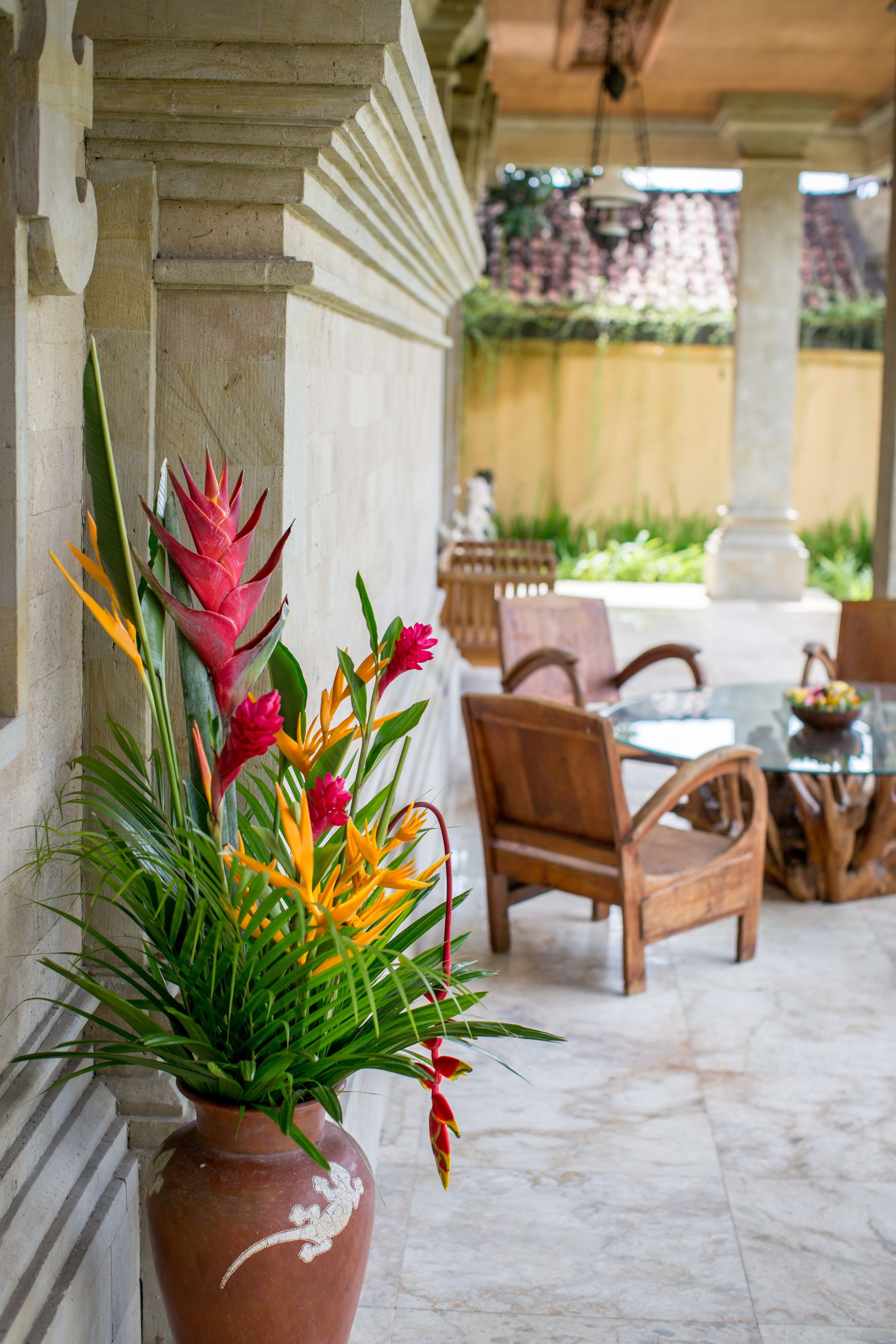 Un espace patio agrémenté d’un grand vase en terre cuite contenant des fleurs tropicales, de chaises en bois et d’une table en verre.