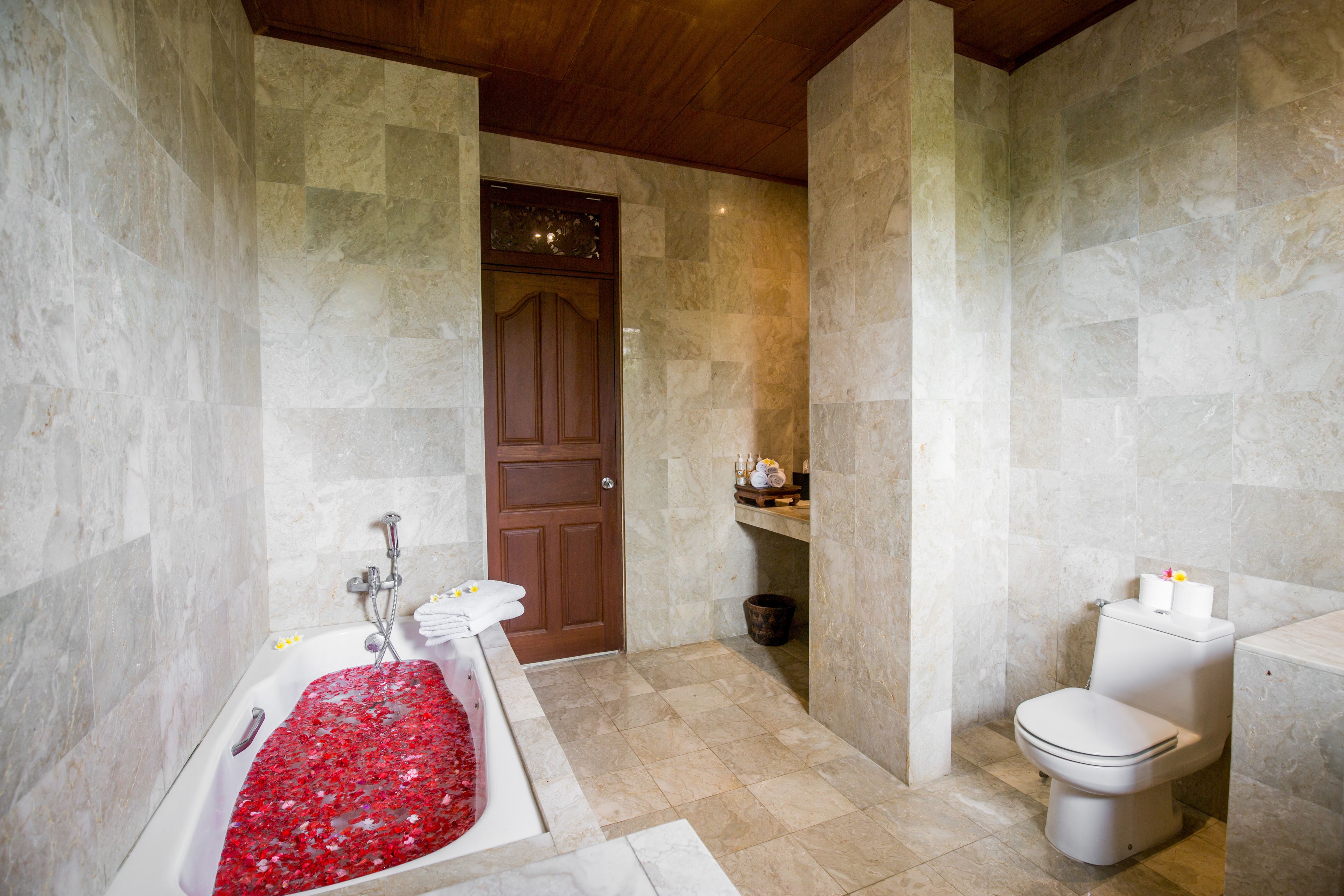 Une salle de bain avec un sol en marbre, une baignoire agrémentée de tapis de bain à motifs floraux rouges, une porte en bois et des toilettes.