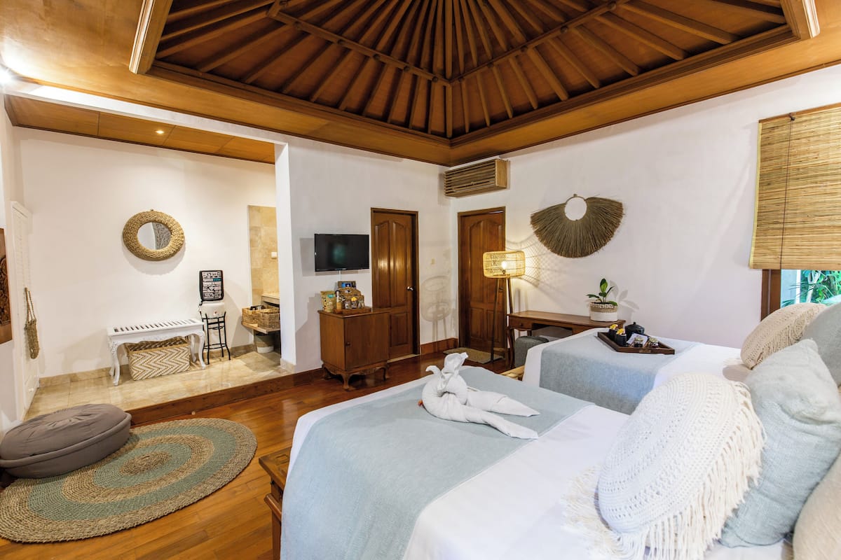 Una habitación de hotel con techo de madera, una cama con colcha blanca y azul, un sofá gris, una mesita con lámpara, un televisor y un mueble de madera.