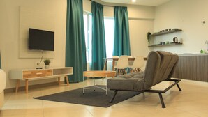Un salon moderne comprenant un canapé, un téléviseur fixé au mur, une table à manger avec des chaises et un coin cuisine avec des étagères.