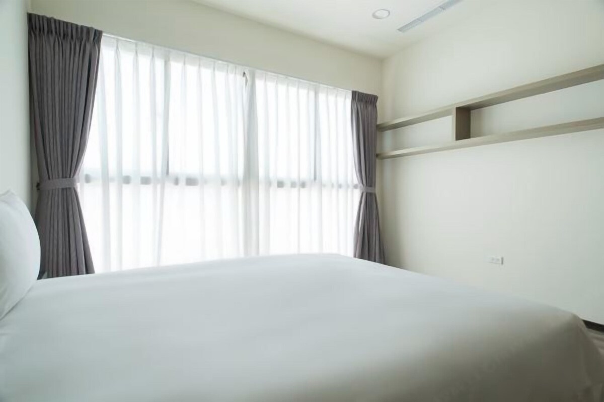 A hotel room with a large bed, a window with curtains, and a built-in shelf above the bed.