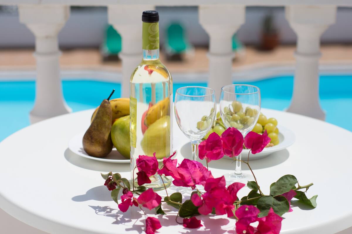 Een tafel met een fles, glazen, fruit en bloemen bij een zwembad.