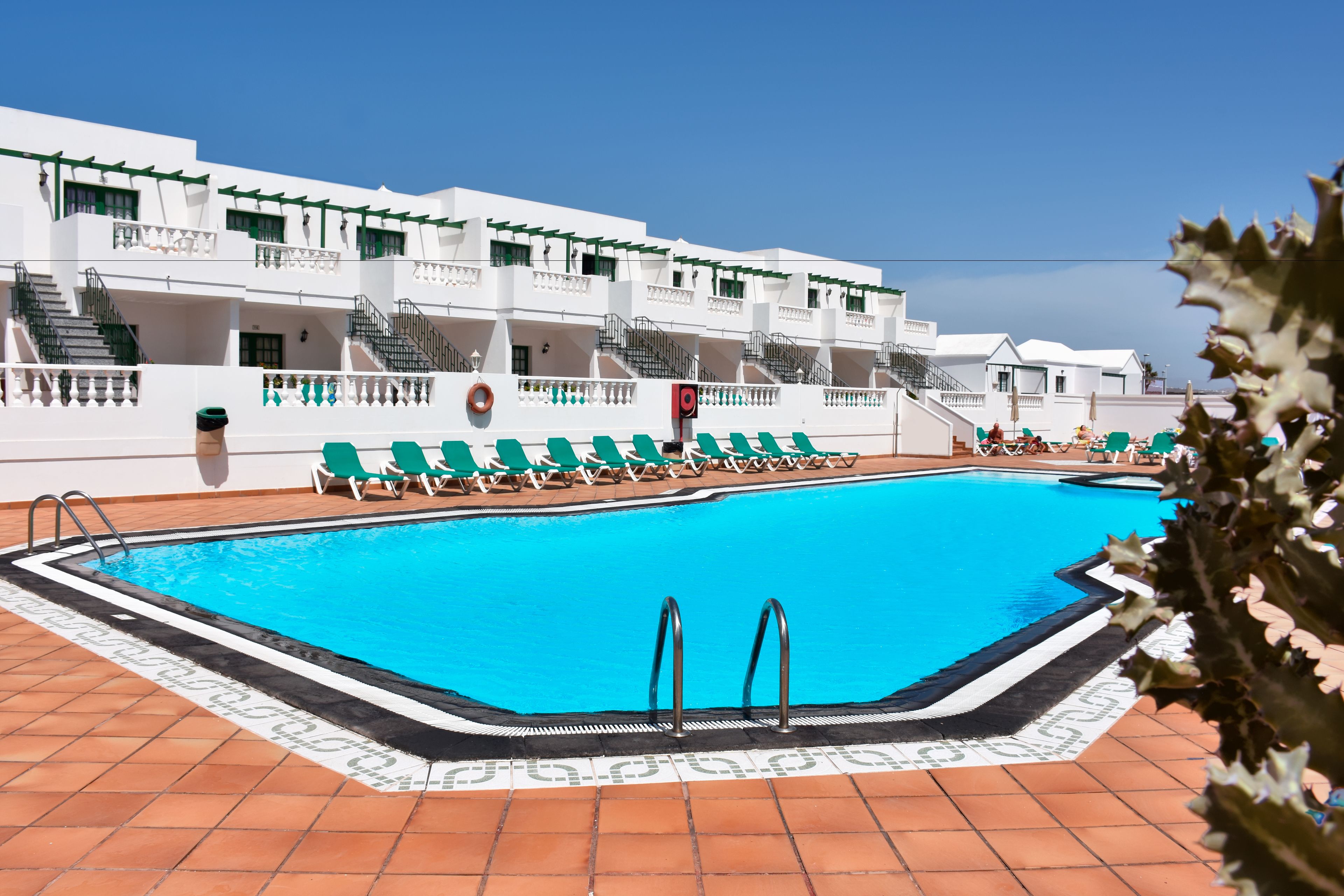 A swimming pool with a tiled deck, green lounge chairs, and a white building with balconies.