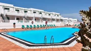 A swimming pool with a tiled deck, green lounge chairs, and a white building with balconies.