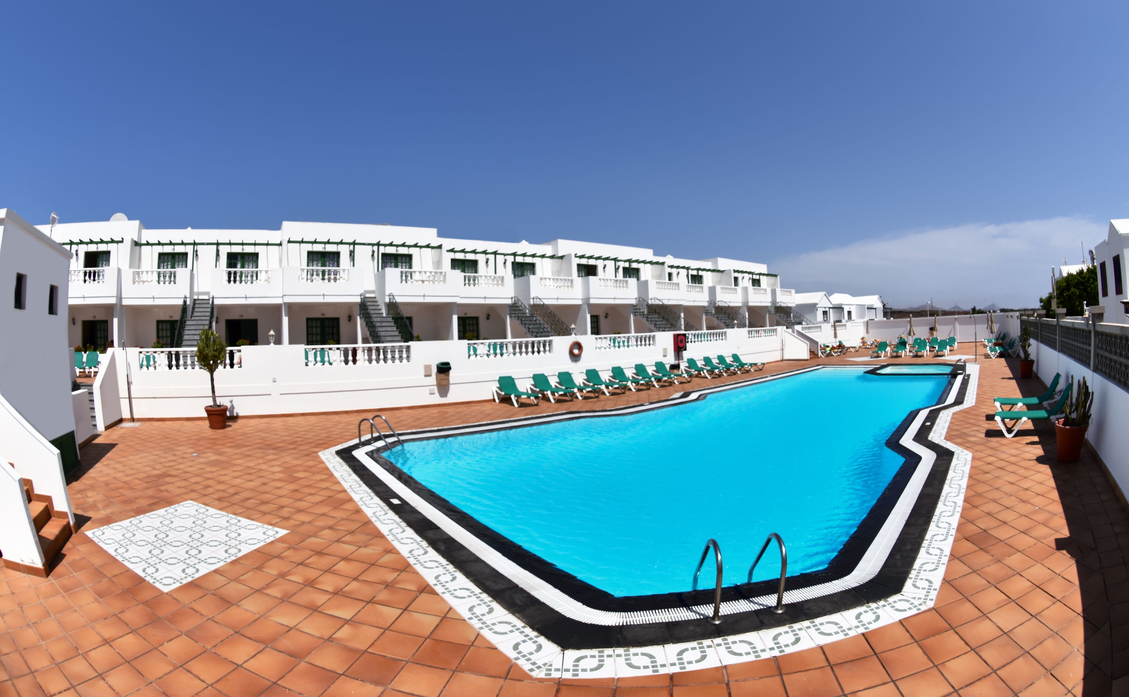 Une piscine sur le toit, avec une eau d’un bleu limpide, entourée de bâtiments blancs dotés de balcons et d’un sol carrelé.