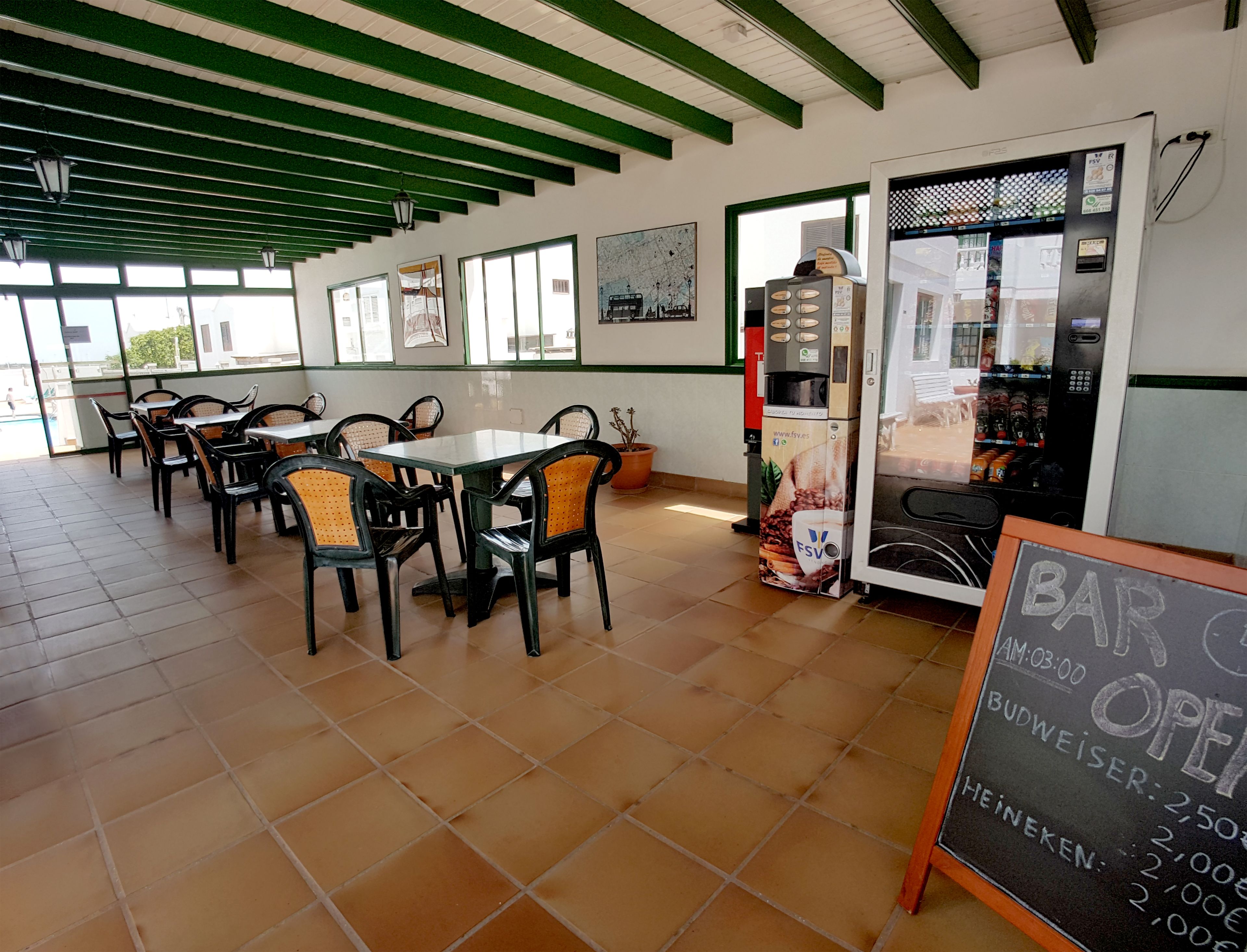A café with outdoor seating, a coffee machine, and a chalkboard sign indicating the opening time and prices for Budweiser and Heineken.