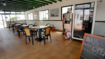 A café with outdoor seating, a coffee machine, and a chalkboard sign indicating the opening time and prices for Budweiser and Heineken.