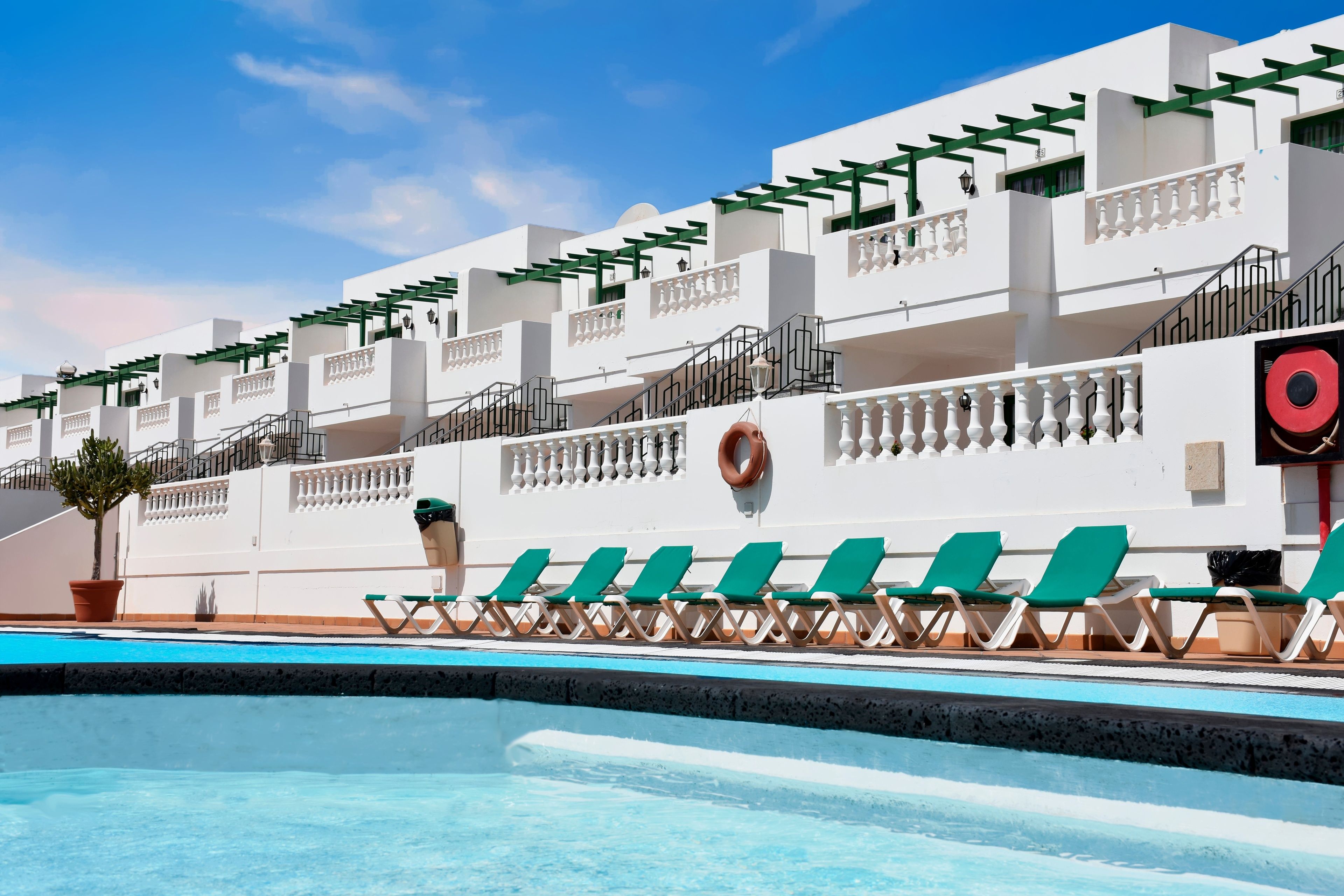 A pool area with green lounge chairs, a lifebuoy, and a white building with balconies.