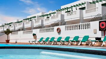 A pool area with green lounge chairs, a lifebuoy, and a white building with balconies.